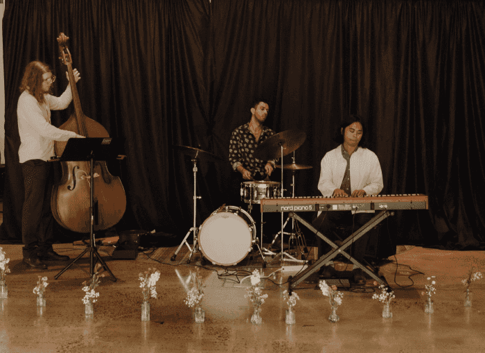 The David Caalman Trio performing live, featuring a bassist, drummer, and pianist in front of a black curtain, with small flower vases arranged on the floor.